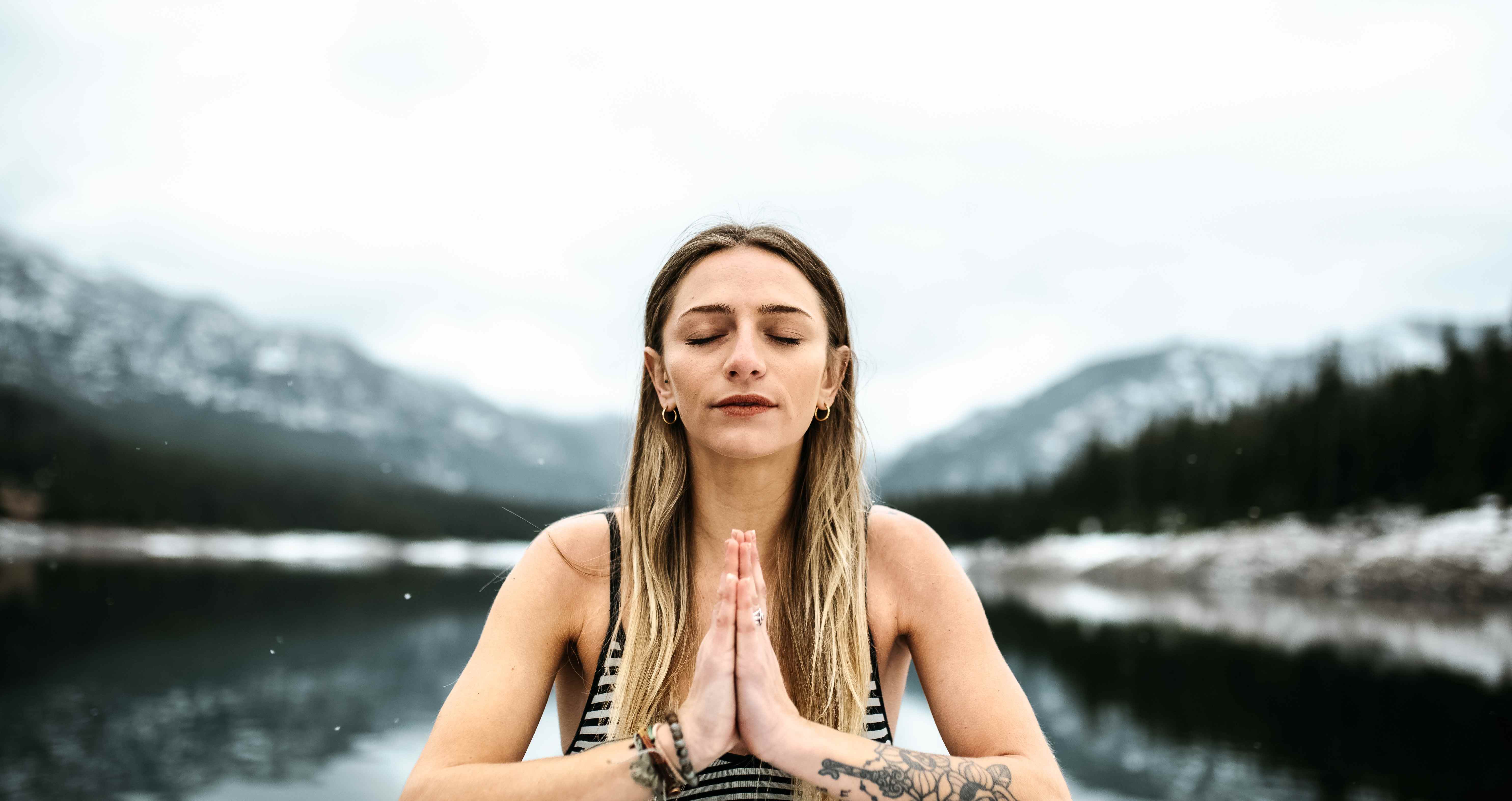 A woman meditating with hands pressed together in front of a mountain lake